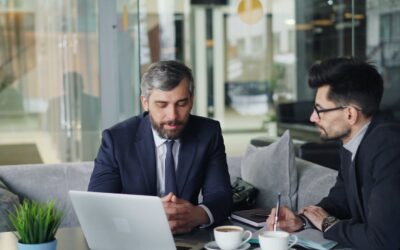 Two businessmen in suits discussing work at a cafe with laptops and coffee.